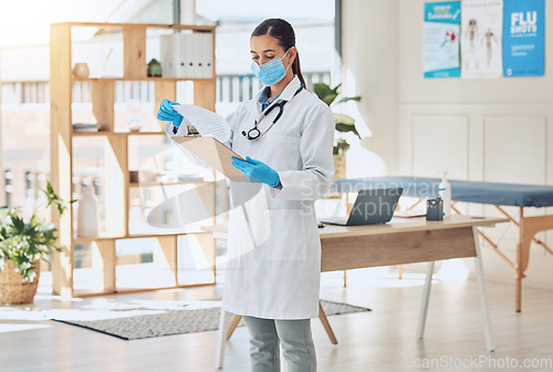 Image of Medical doctor with a clipboard and face mask in her office reading covid results during pandemic. Young healthcare employee doing hospital paperwork in the clinic room in a medicare surgery center.