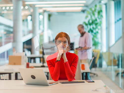 Image of In a modern startup office, a professional businesswoman with orange hair sitting at her laptop, epitomizing innovation and productivity in her contemporary workspace