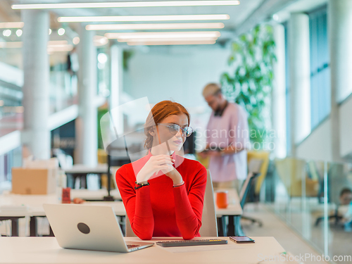 Image of In a modern startup office, a professional businesswoman with orange hair sitting at her laptop, epitomizing innovation and productivity in her contemporary workspace