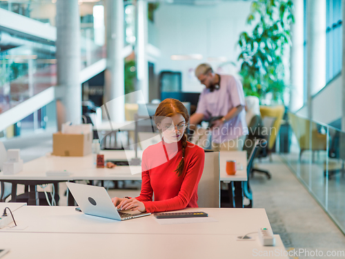 Image of In a modern startup office, a professional businesswoman with orange hair sitting at her laptop, epitomizing innovation and productivity in her contemporary workspace