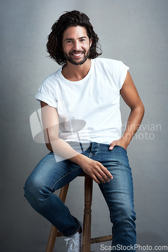 Image of Fashion, smile and portrait of man in studio on a stool with casual, cool and stylish outfit. Happy, handsome and confident young male model from Mexico with trendy style on chair by gray background.