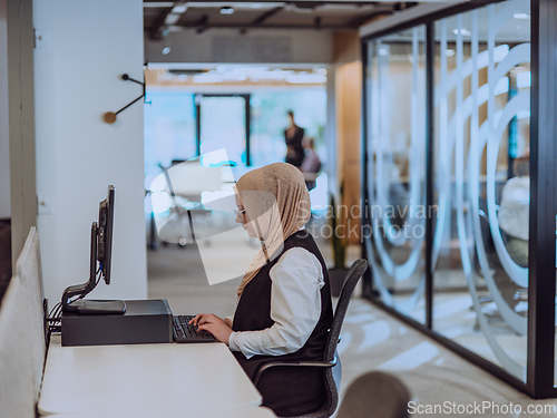 Image of In a modern office, a young Muslim entrepreneur wearing a hijab sits confidently and diligently works on her computer, embodying determination, creativity, and empowerment in the business world