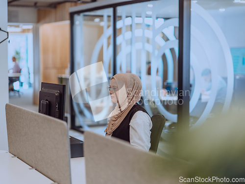 Image of In a modern office, a young Muslim entrepreneur wearing a hijab sits confidently and diligently works on her computer, embodying determination, creativity, and empowerment in the business world