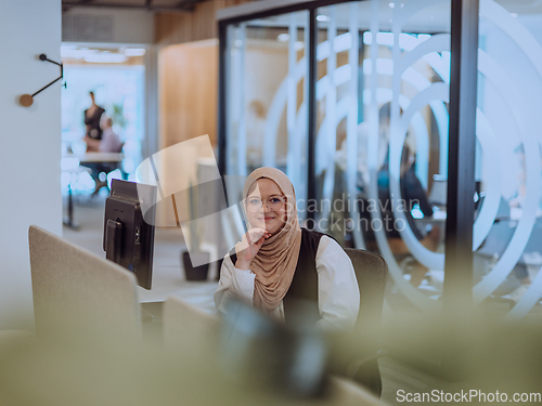 Image of In a modern office, a young Muslim entrepreneur wearing a hijab sits confidently and diligently works on her computer, embodying determination, creativity, and empowerment in the business world