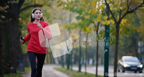 Image of Young beautiful woman running in autumn park and listening to music with headphones on smartphone