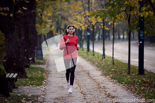 Image of Young beautiful woman running in autumn park and listening to music with headphones on smartphone