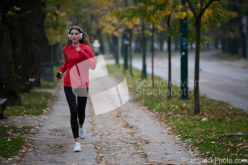 Image of Young beautiful woman running in autumn park and listening to music with headphones on smartphone