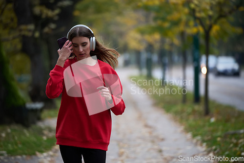 Image of Young beautiful woman running in autumn park and listening to music with headphones on smartphone