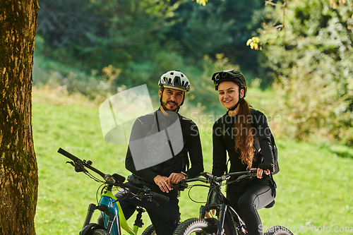 Image of A blissful couple, adorned in professional cycling gear, enjoys a romantic bicycle ride through a park, surrounded by modern natural attractions, radiating love and happiness