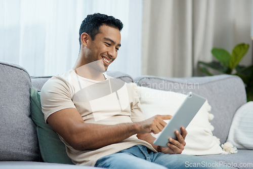 Image of Tablet, smile and relax with a man on a sofa in the living room of his home for social media browsing. Technology, app or gaming search with a happy young person in his apartment on the weekend