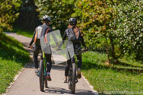 Image of A blissful couple, adorned in professional cycling gear, enjoys a romantic bicycle ride through a park, surrounded by modern natural attractions, radiating love and happiness