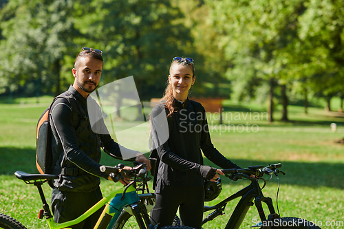 Image of A blissful couple, adorned in professional cycling gear, enjoys a romantic bicycle ride through a park, surrounded by modern natural attractions, radiating love and happiness