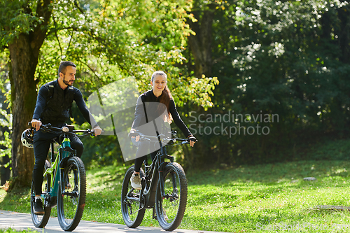 Image of A blissful couple, adorned in professional cycling gear, enjoys a romantic bicycle ride through a park, surrounded by modern natural attractions, radiating love and happiness