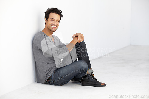 Image of Smile, fashion and portrait of young man by a white wall in empty room with casual, cool and trendy outfit. Happy, confidence and handsome male model from Canada sitting on floor with edgy style.