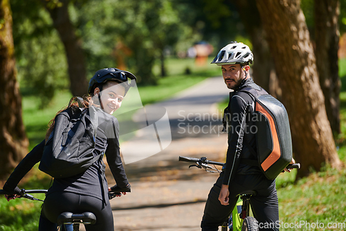 Image of A blissful couple, adorned in professional cycling gear, enjoys a romantic bicycle ride through a park, surrounded by modern natural attractions, radiating love and happiness