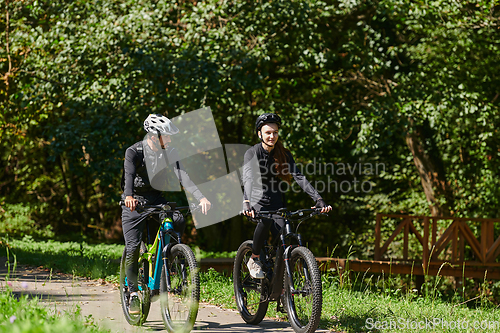 Image of A blissful couple, adorned in professional cycling gear, enjoys a romantic bicycle ride through a park, surrounded by modern natural attractions, radiating love and happiness