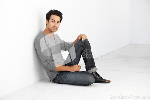 Image of Serious, fashion and portrait of young man by white wall with casual, stylish and trendy outfit. Attractive, confidence and handsome male model from Canada sitting on floor with edgy and cool style.