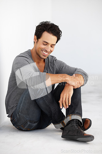 Image of Smile, fashion and handsome young man by white wall with casual, cool and trendy outfit. Happy, confident and attractive male model from Canada sitting on floor with edgy style and positive attitude.