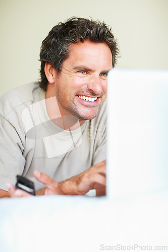 Image of Relax, phone and laptop with a man in the bedroom of his home for entertainment on weekend time off. Smile, tech and a happy man lying in bed, streaming a movie or series while internet browsing