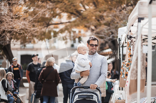 Image of Father walking carrying his infant baby boy child and pushing stroller in crowd of people visiting Sunday flea market in Malaga, Spain.
