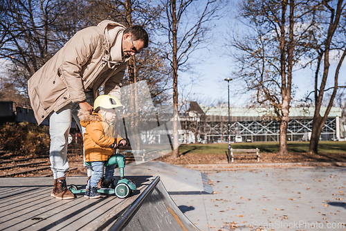 Image of Father supervises his fearless small toddler boy while riding baby scooter outdoors in urban skate park. Child wearing yellow protective helmet
