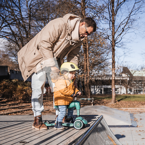 Image of Father supervises his fearless small toddler boy while riding baby scooter outdoors in urban skate park. Child wearing yellow protective helmet