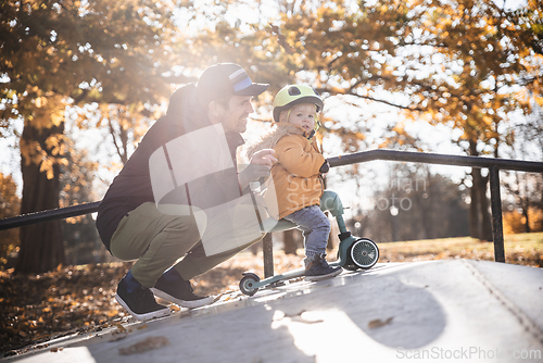 Image of Father supervises his fearless small toddler boy while riding baby scooter outdoors in urban skate park. Child wearing yellow protective helmet