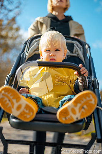 Image of Little toddler child wearing yellow rain coat and rain boots being pushed in stroller by her mother in city park on a sunny autumn day after the rains were gone.