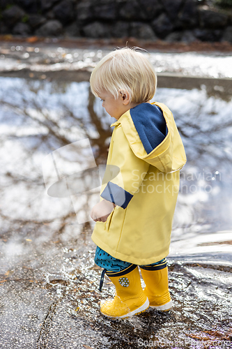 Image of Small blond infant boy wearing yellow rubber boots and yellow waterproof raincoat walking in puddles on a overcast rainy day. Child in the rain.