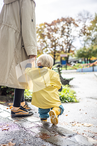 Image of Small bond infant boy wearing yellow rubber boots and yellow waterproof raincoat walking in puddles on a overcast rainy day holding her mother's hand. Mom with small child in rain outdoors.