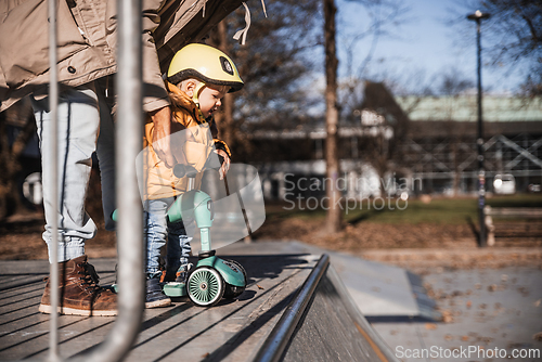 Image of Father supervises his fearless small toddler boy while riding baby scooter outdoors in urban skate park. Child wearing yellow protective helmet