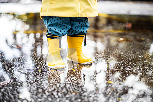 Image of Small infant boy wearing yellow rubber boots and yellow waterproof raincoat standing in puddle on a overcast rainy day. Child in the rain.