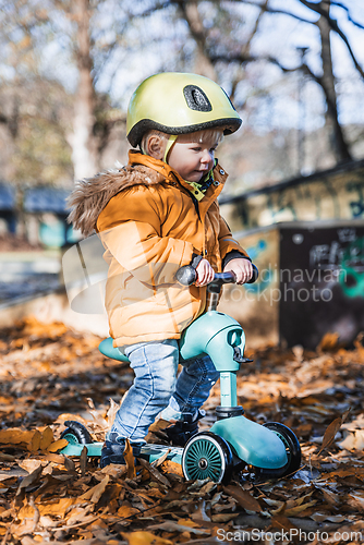 Image of Adorable toddler boy wearing yellow protective helmet riding baby scooter outdoors on autumn day. Kid training balance on mini bike in city park. Fun autumn outdoor activity for small kids.
