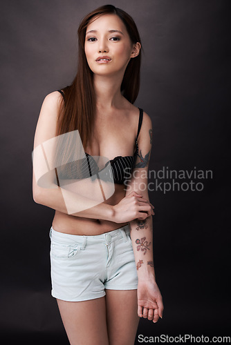 Image of Asian, woman and serious portrait with tattoos in underwear on dark background of studio. Japanese, model and art with ink on body with lady in jeans, shorts and bra to show unique style or skin