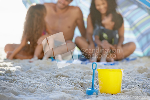 Image of Family, sand and relax on beach with bucket, spade and umbrella for playing by ocean coast. Blue and yellow toy on sandy shore with people in fun bonding, shade or outdoor holiday weekend together