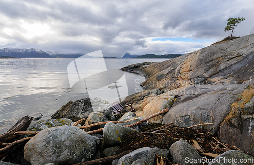 Image of Rocky shore with driftwood and a a chair that has drifted ashore facing the tranquil sea with mountains beyond.