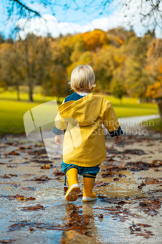 Image of Sun always shines after the rain. Small bond infant boy wearing yellow rubber boots and yellow waterproof raincoat walking in puddles in city park on sunny rainy day.