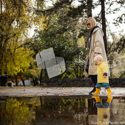 Image of Small bond infant boy wearing yellow rubber boots and yellow waterproof raincoat walking in puddles on a overcast rainy day holding her mother's hand. Mom with small child in rain outdoors.