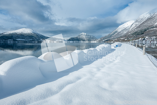 Image of A serene snowy landscape showcases a pathway bordered by undisturbed snow drifts with mountains reflected in the calm sea.