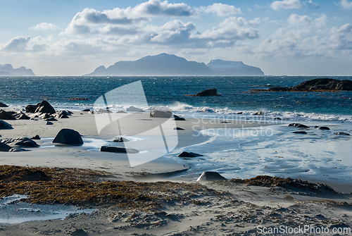 Image of Calm waters wash over a sandy beach with rocks and seaweed, island silhouette in the distance.