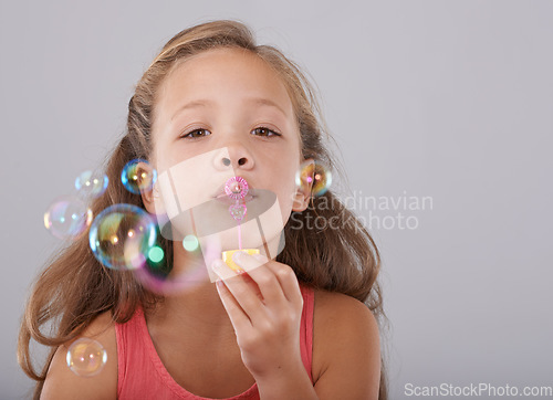 Image of Portrait, little child and blowing bubbles in studio by toy, cute and fun games with soap liquid. Girl, face and learning to play with bubble wand, childhood development and sweet by gray background