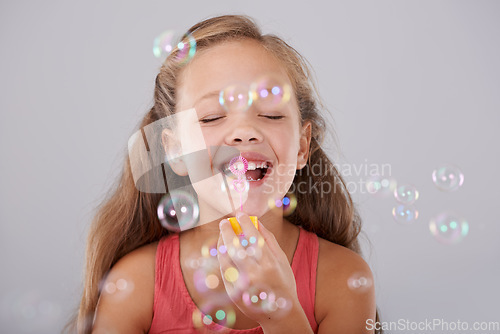 Image of Happy, little girl and blowing bubbles in studio by toy, cute and fun games for hand eye coordination. Child, eyes closed and play with bubble wand, childhood development and smile by gray background