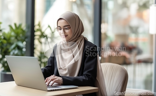 Image of In a cozy cafe, a woman in a hijab embraces modernity as she diligently works on her laptop, seamlessly blending technology with tradition in a contemporary and culturally diverse workspace