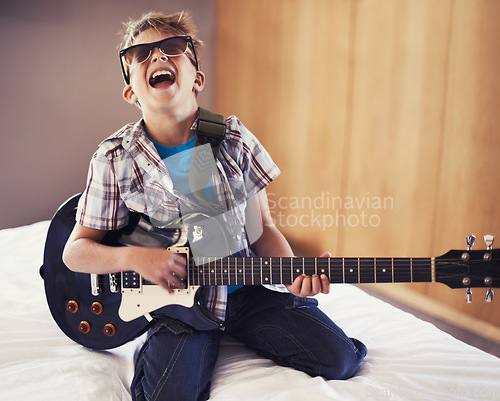 Image of Happy, playing and child with guitar on bed for music lesson with sunglasses at modern home. Fun, rockstar and young boy kid learning with electric string instrument in bedroom for hobby at house.