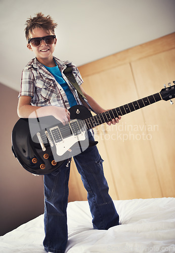 Image of Smile, playing and kid with guitar on bed for music lesson with sunglasses at modern home. Fun, rockstar and young boy child learning with electric string instrument in bedroom for hobby at house.