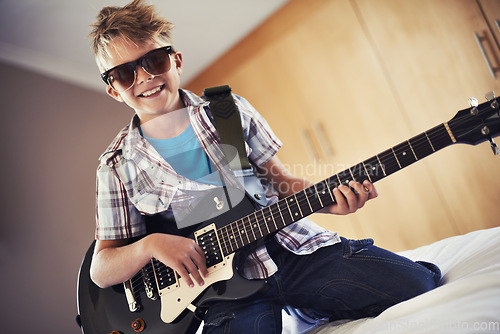 Image of Smile, playing and child with guitar on bed for music lesson with sunglasses at modern home. Fun, rockstar and young boy kid learning with electric string instrument in bedroom for hobby at house.