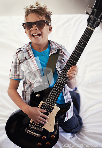 Image of Happy, playing and boy kid with guitar on bed for music lesson with sunglasses at modern home. Fun, rockstar and young child learning with electric string instrument in bedroom for hobby at house.