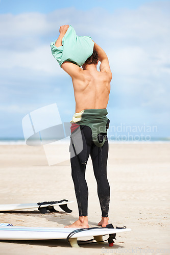 Image of Surfing, man and getting ready with surfboard on beach with wetsuit, blue sky and dressing with mock up space. Rear view, person and athlete by ocean for training, surfer workout and extreme sports