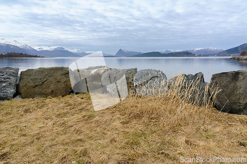 Image of Tranquil scene showing the calm sea, boulders, and mountains under a soft evening sky.