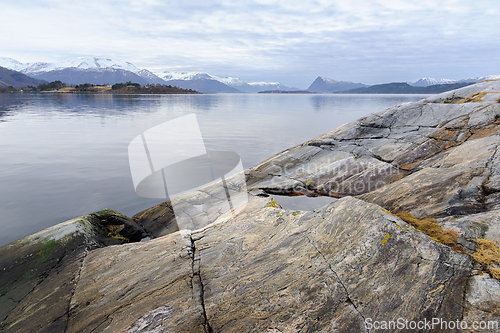 Image of A tranquil seaside setting showing a rocky shore and distant snow-covered mountains under a soft evening sky.